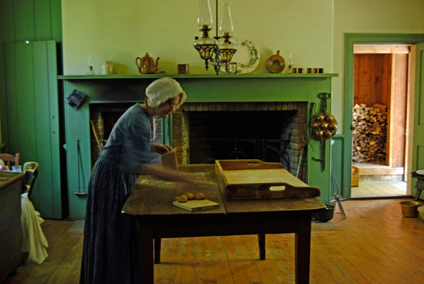 a woman dressed in pioneer clothing works at a kitchen table