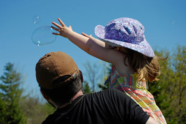 a father holds up a little girl as she tries to reach a floating bubble